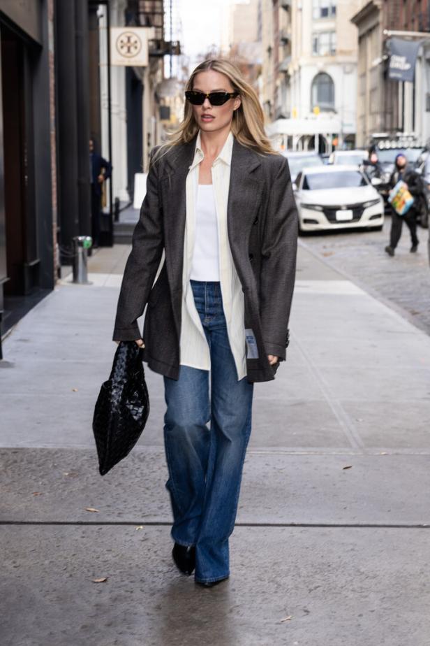 Margot Robbie geht mit Sonnenbrille, Blazer und Handtasche auf dem Bürgersteig entlang.