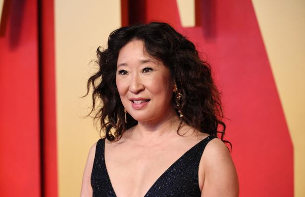 Sandra Oh lächelt vor einem roten und beigen Hintergrund.