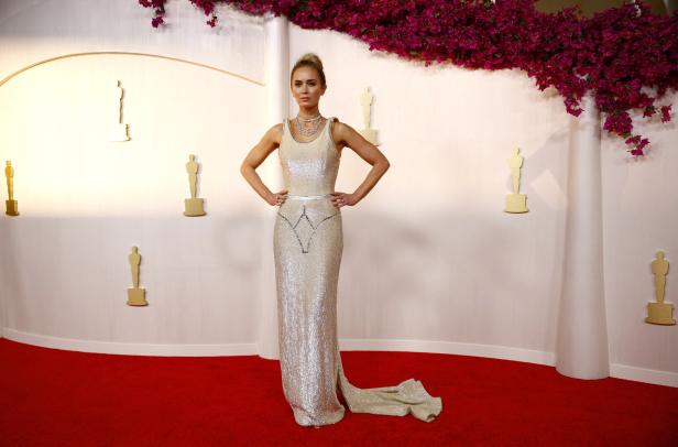 Eine Frau in einem glitzernden Kleid posiert auf dem roten Teppich vor einer Wand mit Oscar-Statuetten.