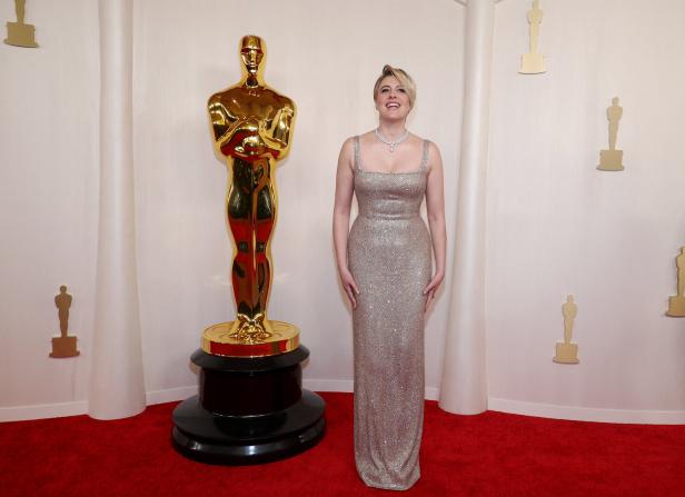 Greta Gerwig posiert auf dem roten Teppich neben einer Oscar-Statue.