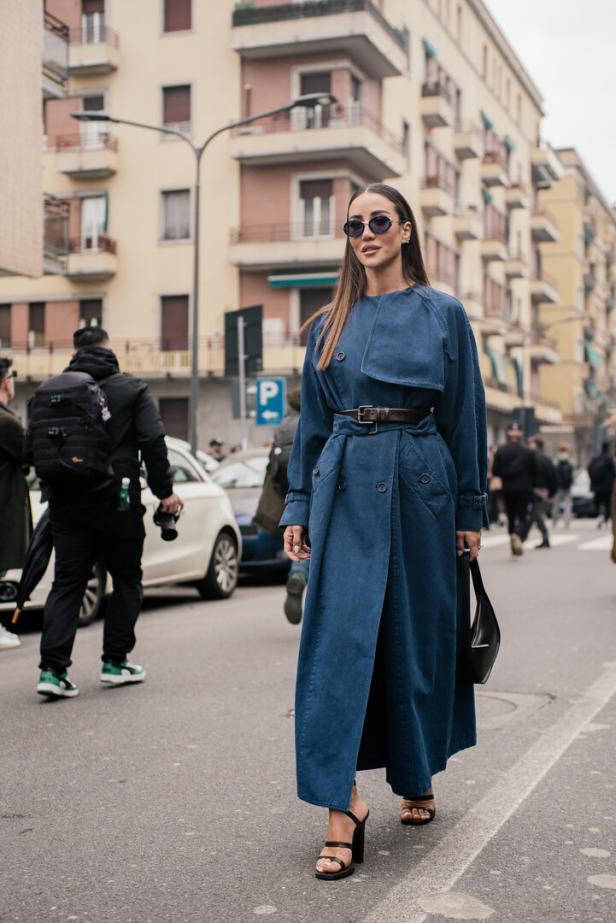 Eine Frau mit Sonnenbrille und blauem Trenchcoat geht eine Straße entlang.