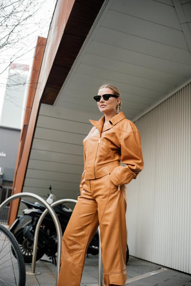 Eine Frau in einem braunen Lederanzug und Sonnenbrille steht im Freien.