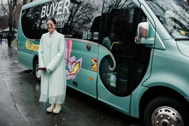 Eine Frau in einem hellgrünen Mantel steht vor einem Bus mit der Aufschrift „Buyer Milan Fashion Week 20-26 2024“.