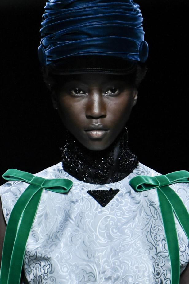Eine Frau mit blauem Hut und einem Kleid mit grünen Schleifen präsentiert ein Prada-Design.
