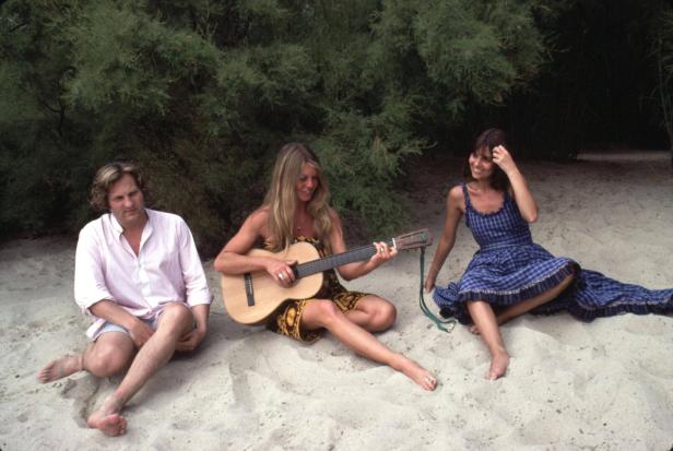 Brigitte Bardot sitzt mit zwei Freunden am Strand, eine Gitarre spielend.