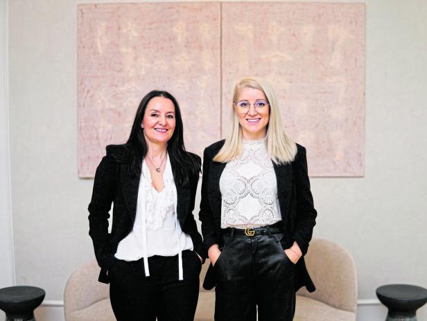 Zwei Frauen posieren in schwarzen Blazern vor einem abstrakten Kunstwerk.
