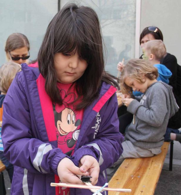 Ein Mädchen bastelt mit Stöcken und einer Klammer, während andere Kinder im Hintergrund sitzen.