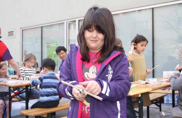 Ein Mädchen bastelt mit Holzstäben, während andere Kinder an Tischen sitzen.