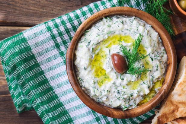 Griechisches Tzatziki in einer Holzschüssel mit Dille, Olivenöl und einer Olive garniert, daneben griechisches Brot und ein grünes Tuch
