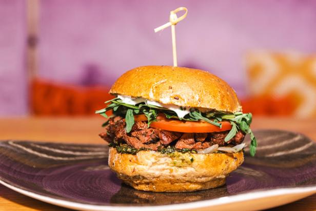 Ein Burger mit Rindfleisch, Tomaten, Rucola und einer hellen Soße wird auf einem Teller serviert.