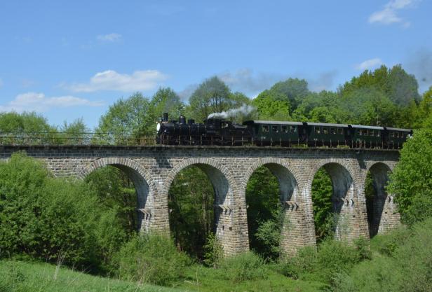 Ein Zug fährt über eine alte Steinbrücke, umgeben von grüner Landschaft und klarem Himmel.