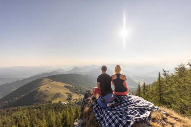 Ein Paar sitzt auf einem Gipfel und blickt in die Ferne über eine bewaldete Hügellandschaft.