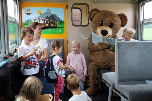Eine Gruppe von Kindern sitzt in einem Zugwaggon und unterhält sich mit einem großen Teddybären.