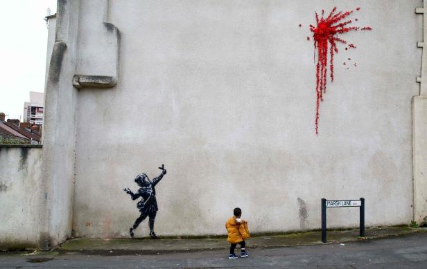 Ein Kind betrachtet ein Banksy-Graffiti an einer Hauswand, bei dem rote Farbe wie von einem Ballon herabläuft.