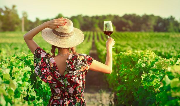 Frau im Sommerkleid und mit Sonnenhut hebt ein Weinglas mit Rotwein hoch, steht inmitten von Weinbergen