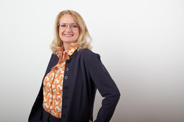 Eine lächelnde Frau mit blonden Haaren, Brille und einer blau-orangenen Bluse vor einem weißen Hintergrund.