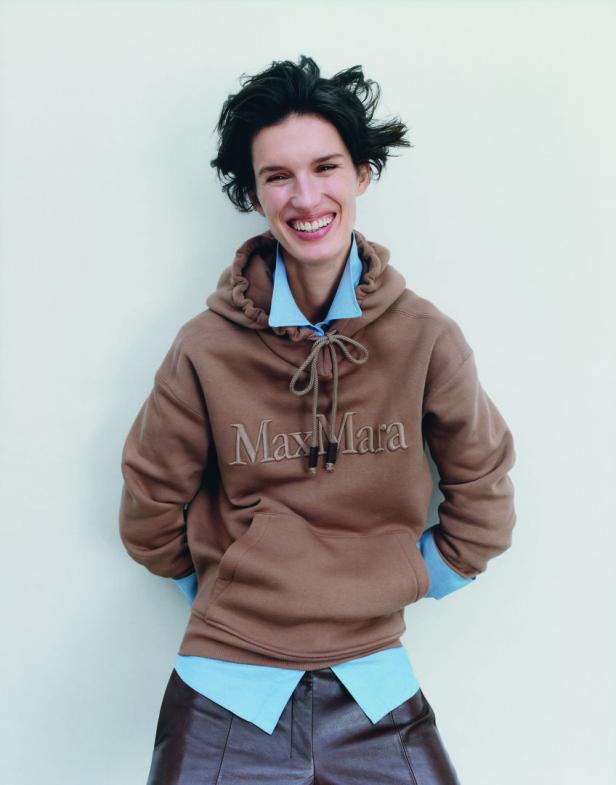 Eine Frau mit braunem Max Mara-Hoodie und Lederhose lächelt.