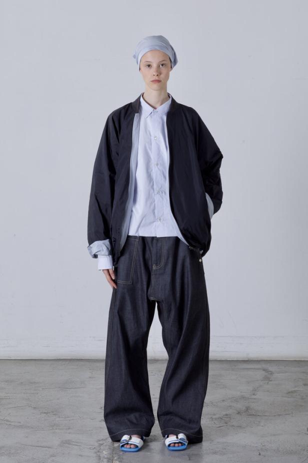 Eine Person in einer Jacke, einem hellblauen Hemd, einer weiten Jeans und Sandalen steht vor einer weißen Wand.