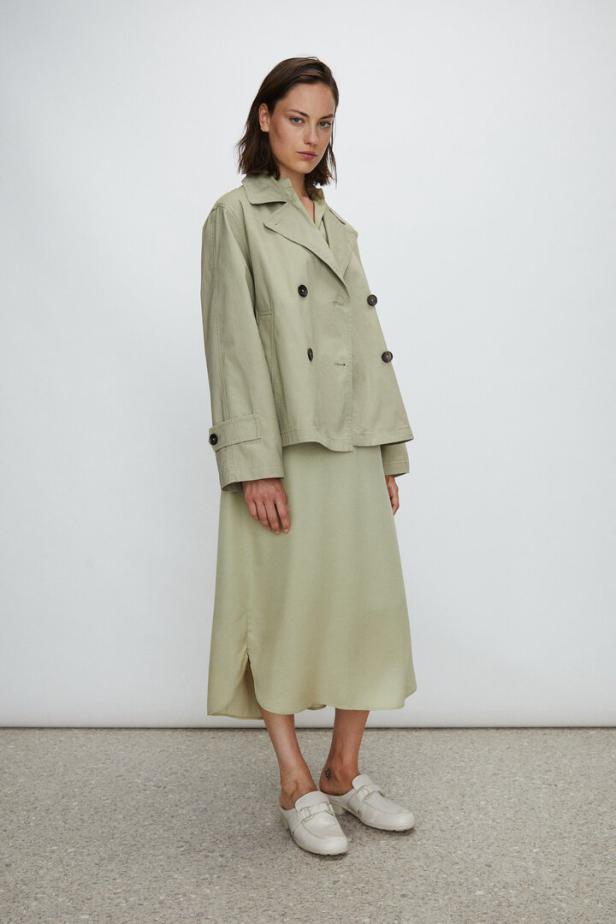 Eine Frau trägt einen hellgrünen Trenchcoat und ein passendes Kleid mit weißen Schuhen.