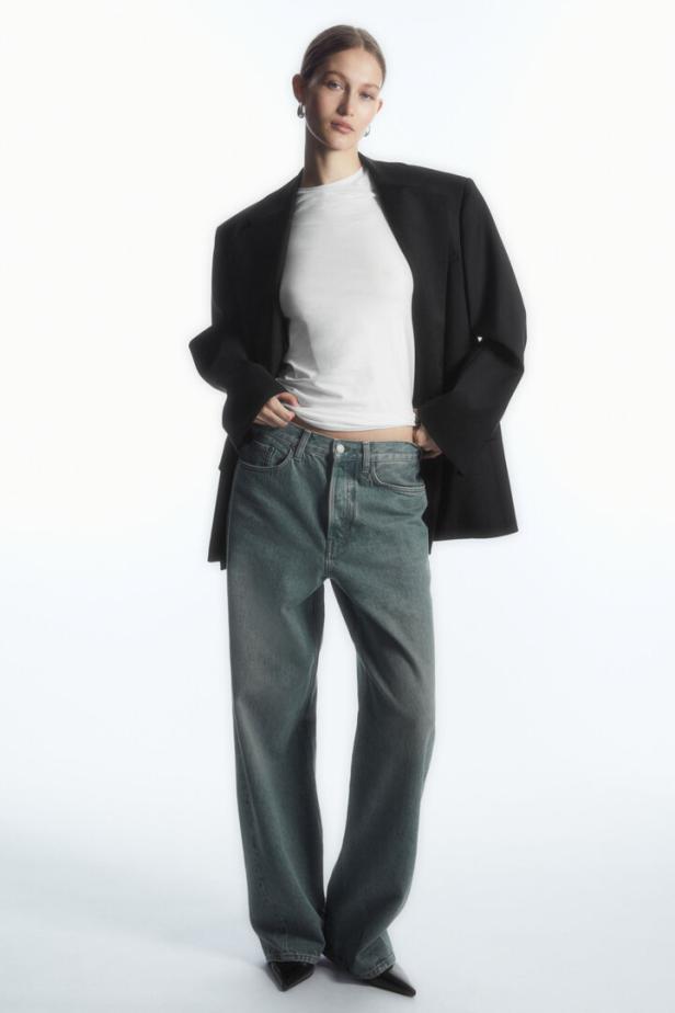 Eine Frau in Jeans, T-Shirt und Blazer posiert vor einem weißen Hintergrund.