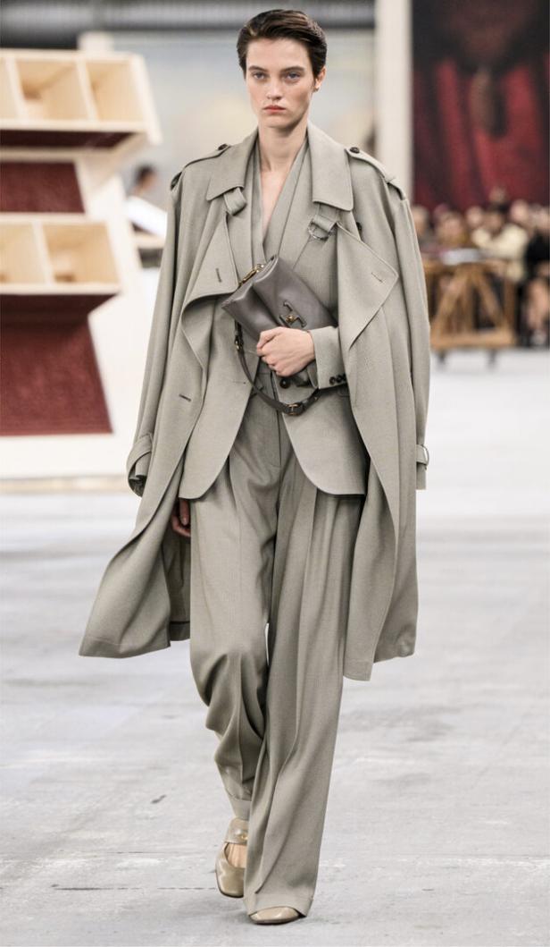 Eine Frau in einem grauen Hosenanzug und Trenchcoat mit einer Handtasche auf einem Laufsteg.