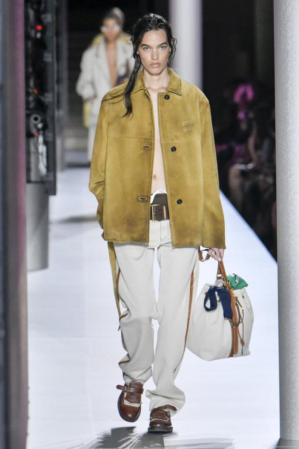 Ein Model auf dem Laufsteg, gekleidet in eine Jacke, helle Hose und braune Schuhe, mit einer Handtasche.