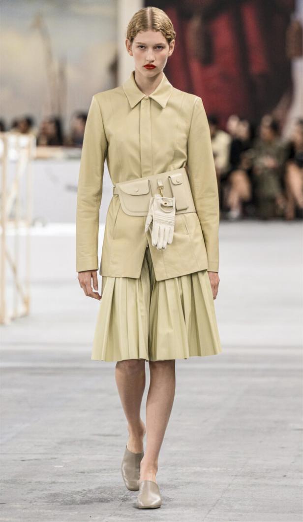 Ein Model präsentiert ein beigefarbenes Ensemble aus Jacke, Rock und Gürteltasche auf einem Laufsteg.