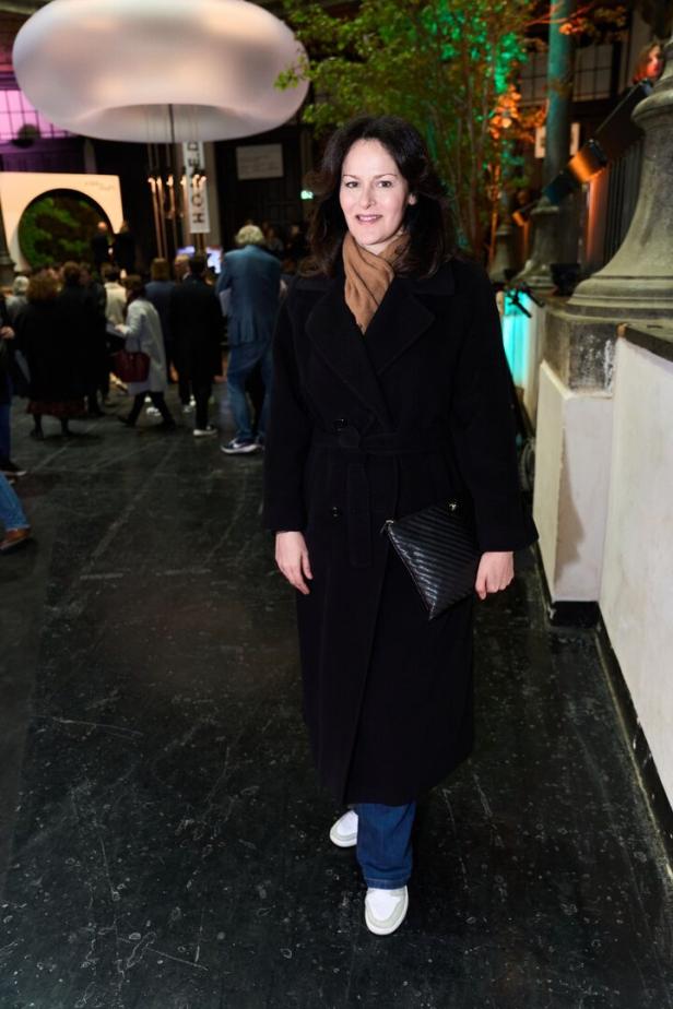 Eine Frau in einem schwarzen Mantel und Jeans steht in einer Halle mit anderen Personen.