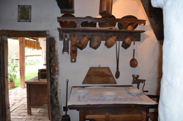 Rustikaler Raum mit Holztisch, Geschirr, alten Küchenutensilien an der Wand und Blick nach draußen auf einen Hof mit Pflanzen.