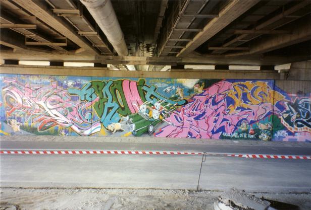 Eine Wand unter einer Brücke ist mit Graffiti und Cartoonfiguren bemalt.
