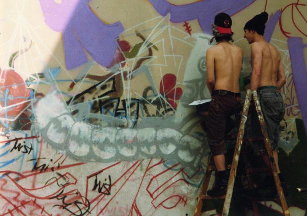 Zwei Männer stehen auf einer Leiter und sprühen Graffiti auf eine Wand.