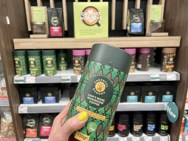 Eine Hand hält eine Dose „Cheerful Buddha Lion's Mane Mushroom Coffee“ vor einem Regal mit Kaffee.