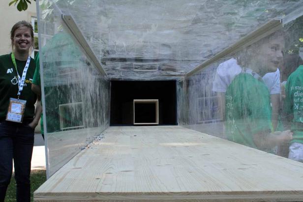 Eine Frau und Kinder stehen vor einem transparenten Tunnel mit einem Würfel am Ende.