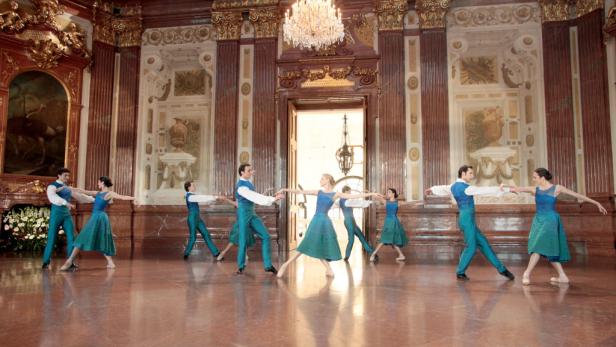Eine Gruppe von Tänzern führt eine Ballettchoreografie in einem prunkvollen Saal auf.