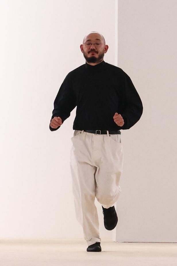 Ein Mann mit Brille, Bart und schwarzem Pullover geht in einem hellen Raum.