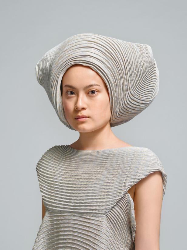 Eine Frau trägt ein plissiertes Kleid und einen passenden Hut in einem futuristischen Design.