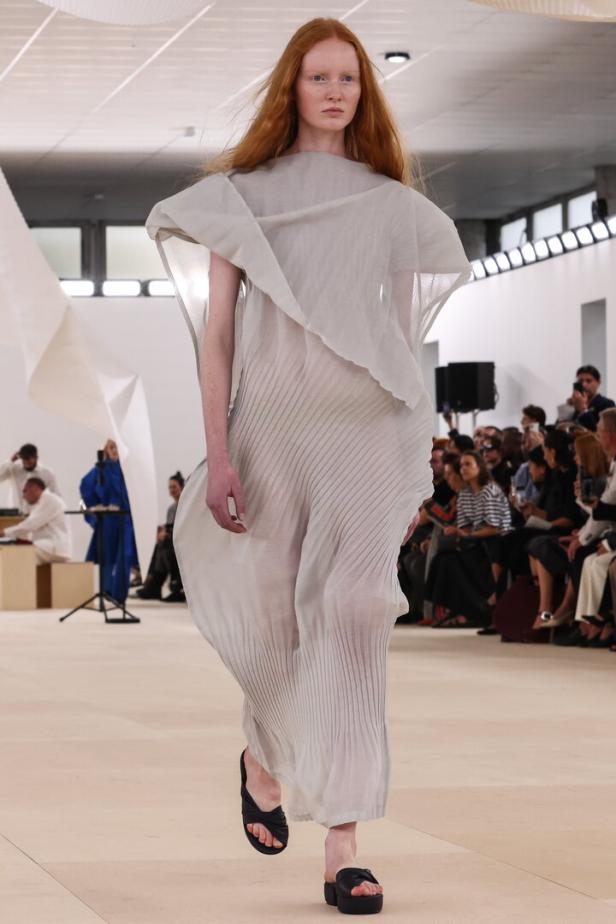 Ein Model präsentiert ein transparentes, plissiertes Kleid und schwarze Sandalen auf dem Laufsteg.