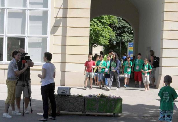 Ein Kamerateam interviewt eine Frau, während eine Gruppe Kinder durch einen Torbogen geht.