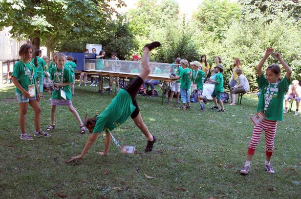 Eine Gruppe Kinder in grünen T-Shirts spielt auf einer Wiese, ein Mädchen macht ein Rad.