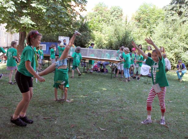 Eine Gruppe Kinder in grünen T-Shirts spielt und turnt auf einer Wiese.