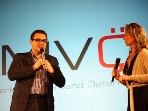Ein Mann und eine Frau stehen mit Mikrofonen vor einem Logo und sprechen.