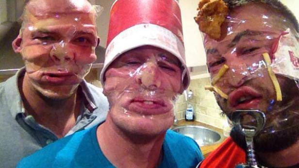 Drei Männer haben Frischhaltefolie um ihre Gesichter gewickelt, einer trägt einen Huteimer, einer hat Pommes und Chicken Nuggets im Gesicht.