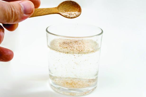 Eine Hand gießt Flohsamenschalen von einem Holzlöffel in ein Glas Wasser.