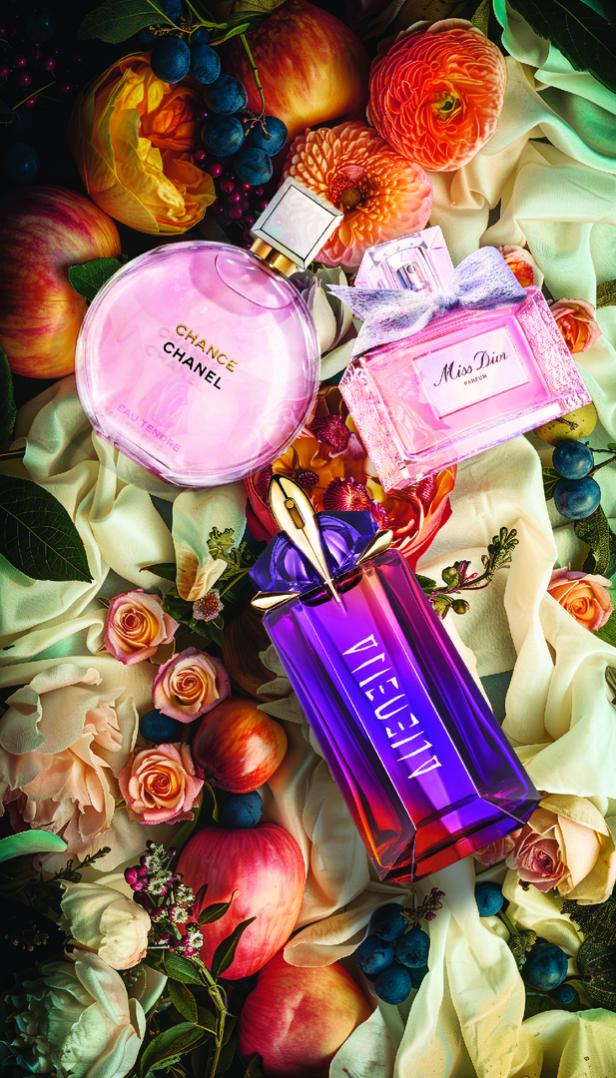 Verschiedene Parfümflaschen von Chanel, Dior und Mugler liegen zwischen Blumen und Früchten auf Stofftüchern.