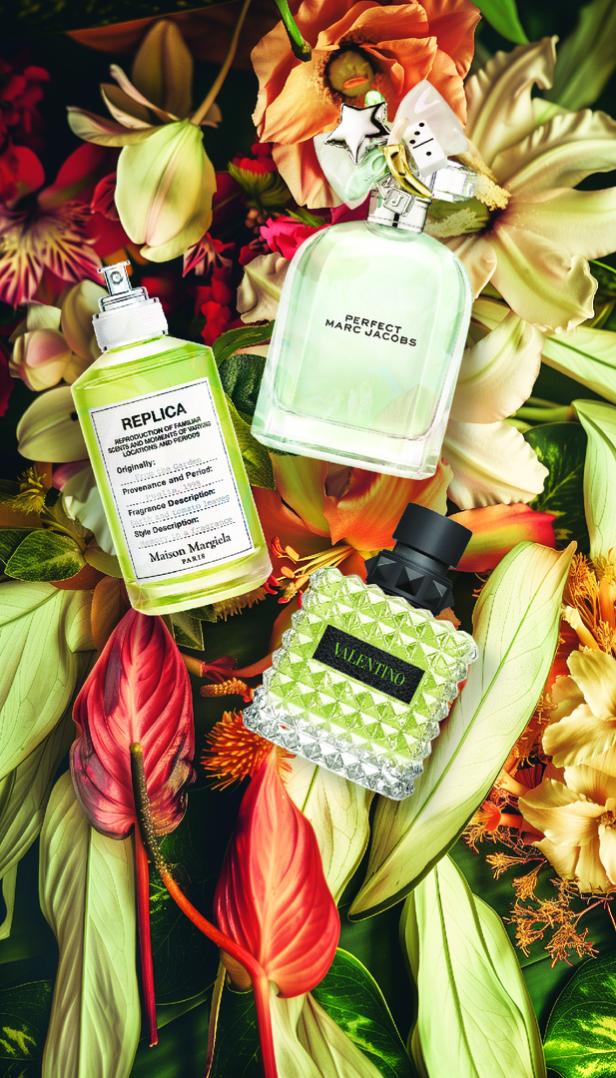 Drei Parfümflaschen von Replica, Marc Jacobs und Valentino liegen zwischen Blumen und Blättern.