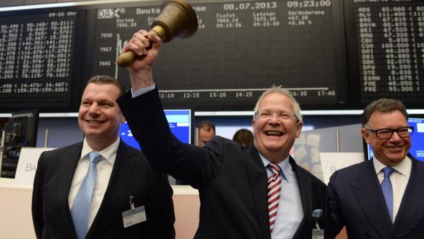Männer läuten die Börsenglocke vor einer Anzeigetafel mit Börsenkursen.