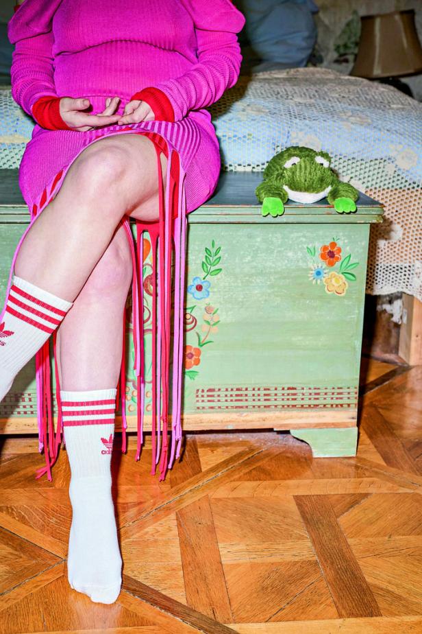 Eine Frau in einem pinkfarbenen Kleid und Adidas-Socken sitzt neben einem grünen Frosch auf einer bemalten Truhe.