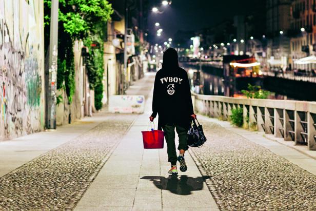 Eine Person mit Kapuzenpullover und Eimer geht nachts eine Gasse entlang.