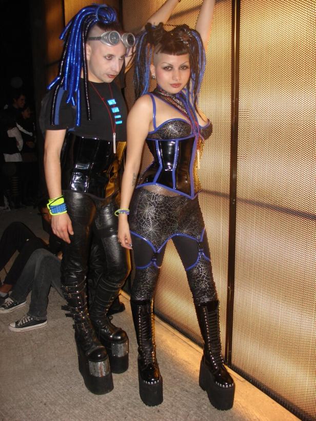 Zwei Personen im Cyberpunk-Stil posieren vor einem Gitter.