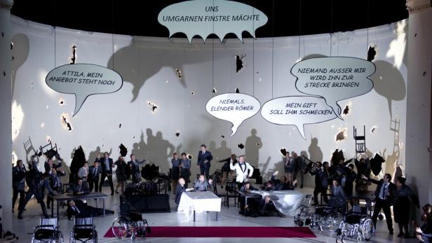 Eine Theaterszene mit Schauspielern, Tischen, Rollstühlen und Sprechblasen an der Wand.